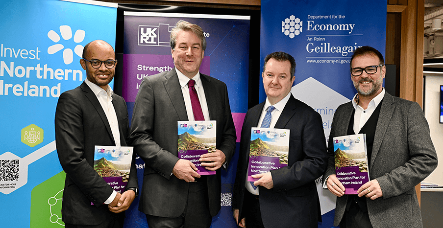Four men standing in front of company pop-up branding, each holding a copy of the Collaborative Innovation plan for Northern Ireland