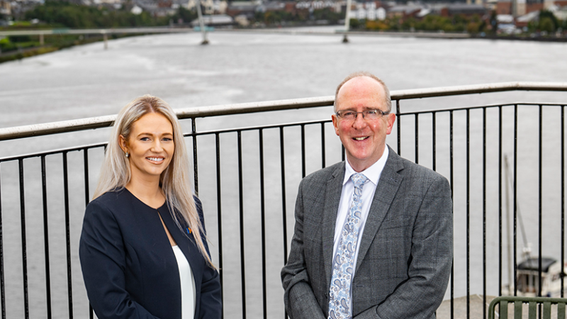 A man and a woman are posing for a photograph on a balcony that overlooks the Peace Bridge 