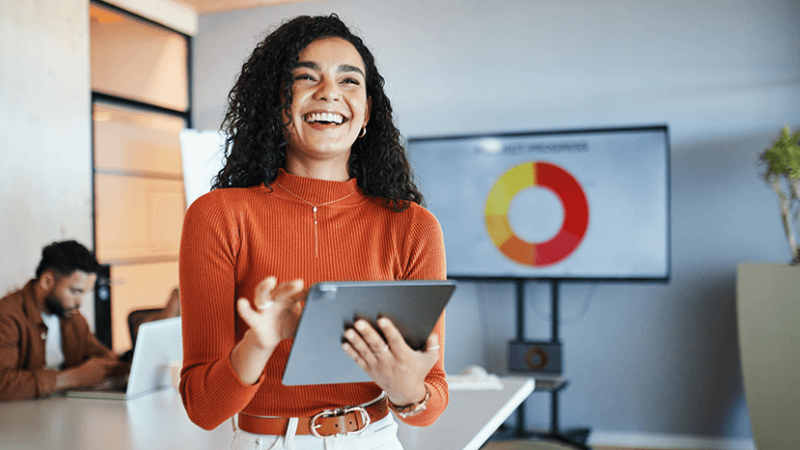 A woman is standing in an office, holding a tablet and smiling